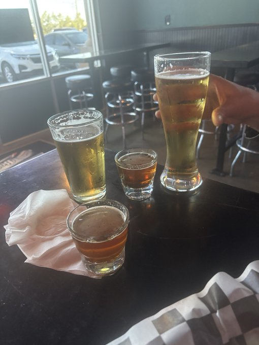 View of four drinks on a dark bar counter including a pilsner glass of beer being held by a hand, alongside a smaller lager glass, and two shot glasses of dark liquor.