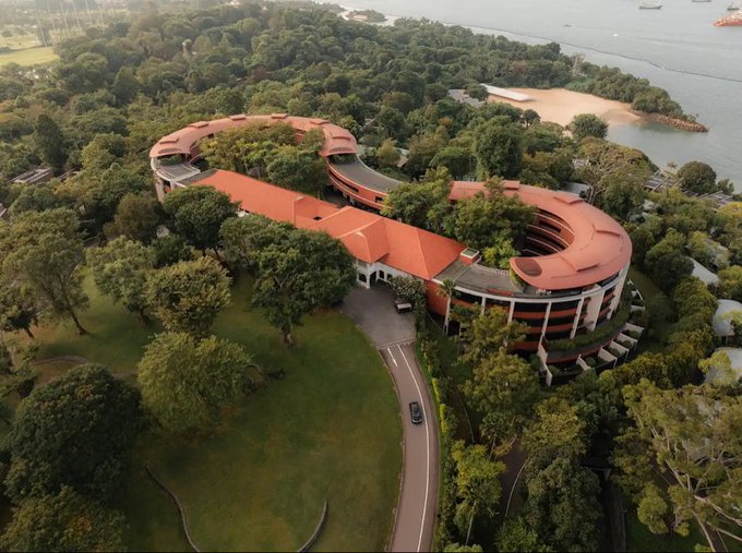 An aerial view shows a large, modern building with distinctive curved red-tiled roofs nestled among lush tropical greenery, with a beach and ships visible in the background—capturing the scenic Singapore setting referenced in the post about an upcoming, highly anticipated Hyperliquid event.