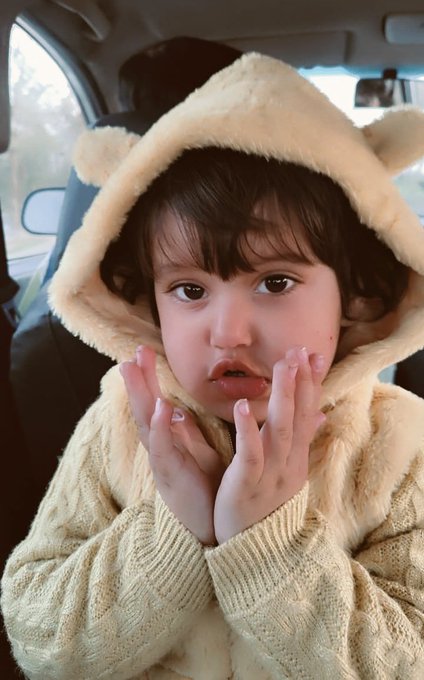 A young child with dark hair wearing a cream-colored bear hooded sweater sits in a car, resting their chin on their hands with a pouty expression.