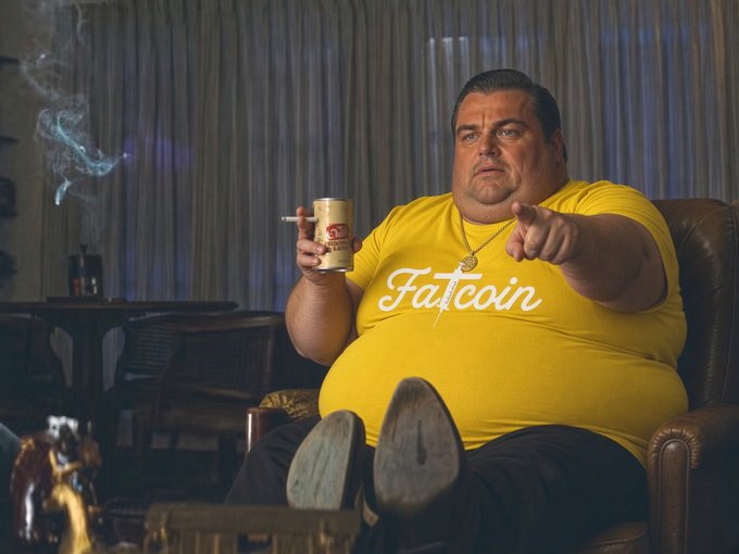 A man wearing a yellow 'Fatcoin' t-shirt sits in a leather chair, smoking a cigarette and holding a can while pointing at the camera.