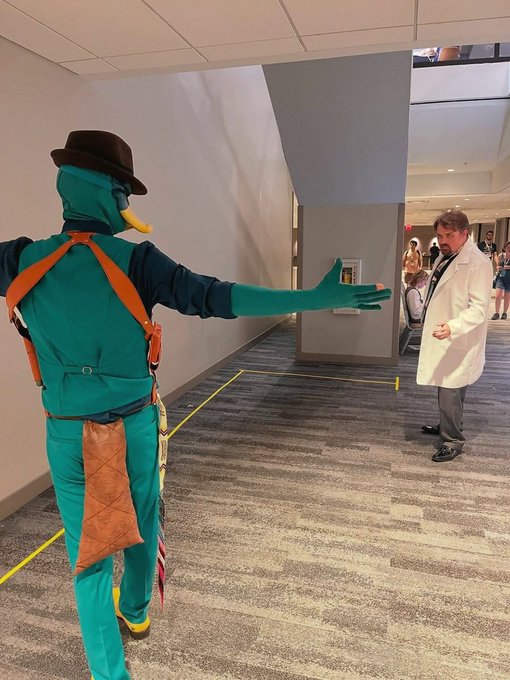 A person in a full green squid-like costume with a brown hat and orange accents holds out their hand in a hallway toward a man wearing a white lab coat.