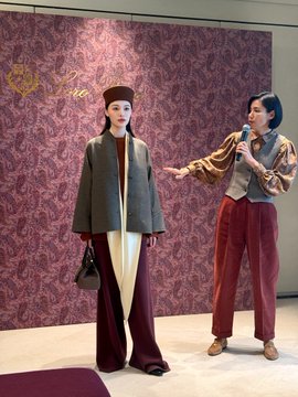 Two women pose in front of a purple damask backdrop; one wears a beret and oversized blazer while the other holds a microphone wearing a vest and maroon pants.