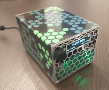 A custom-built computer case with a hexagonal honeycomb pattern that illuminates in green sits on a wooden surface, featuring a 7.032 TRHz display on the front.