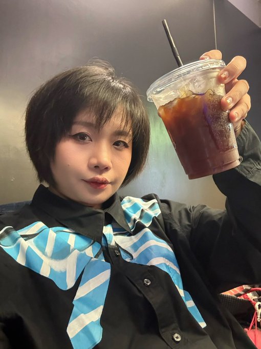 A person with short hair wearing a blue and white striped ruffled shirt holds up a clear plastic cup of iced coffee.