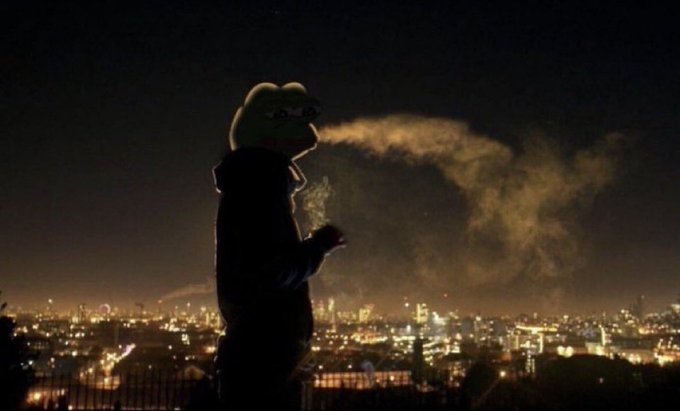 A nighttime photograph showing the silhouette of a person wearing a frog-shaped hood exhaling visible breath vapor, with a city skyline illuminated in the background.