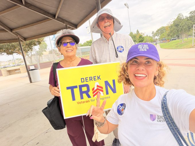 A selfie of three people at a political event holding a yellow campaign sign that reads 'DEREK TRAN - Veteran for Congress', with one person wearing a purple 'GET OUT THE VOTE 2020' cap and another in a 'WALKS FOR CRYPTO' shirt.