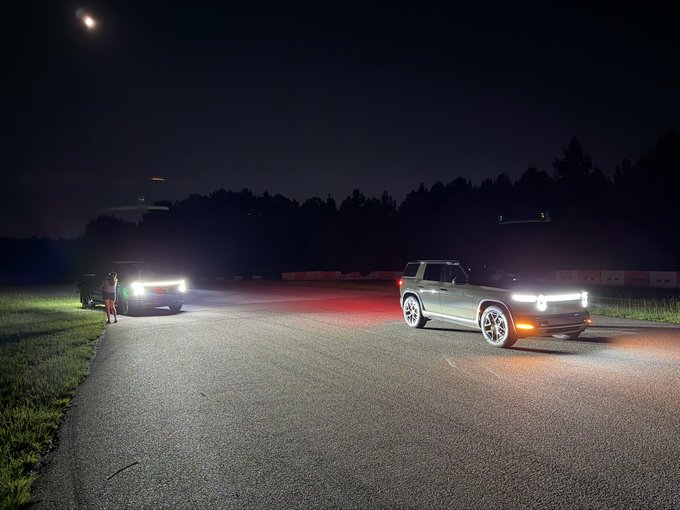 Two SUVs face each other on a dark road at night, their headlights and red brake lights illuminating the pavement, with a person standing near the vehicle on the left.