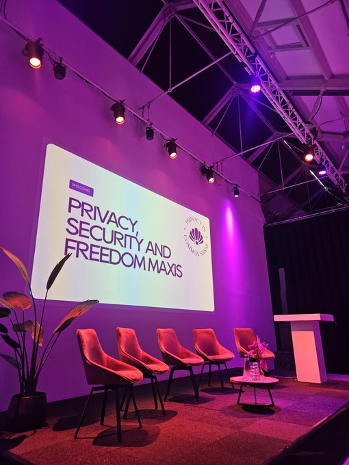 An empty event stage set up for a panel discussion, illuminated with purple lighting and a large screen displaying 'PRIVACY, SECURITY AND FREEDOM MAXIS' with a 'Privacy and Freedom Summit' logo.