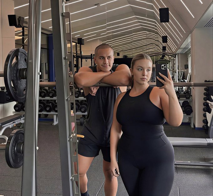 A man and woman pose together in a gym for a mirror selfie, both wearing black athletic outfits, with the woman taking the photo with her phone.

The image directly visualizes the post's joke by showing a fit couple in the gym, humorously suggesting that approaching a "fat girl at the gym" is an undervalued investment opportunity—just like buying Bitcoin early.