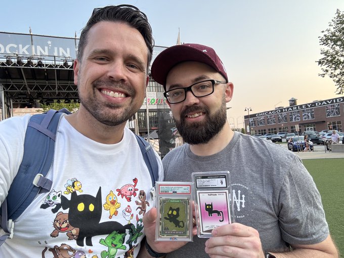 Two smiling men pose together outdoors, one wearing a cat-themed t-shirt and the other holding two PSA-graded trading cards with cat designs, with a Chicago Harley-Davidson sign visible in the background.
