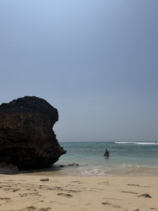 A sunny beach scene with a large rock formation on the left and a person in the water in the distance.