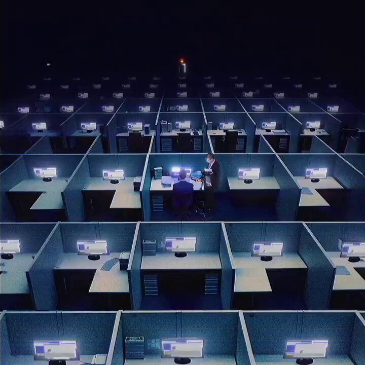 The image shows a large office space filled with rows upon rows of cubicles, each containing multiple computer monitors. Two men are standing at their desks within this workspace, surrounded by numerous computers. The scene appears to be a depiction of an office environment where employees work on their tasks using the provided computers and equipment.