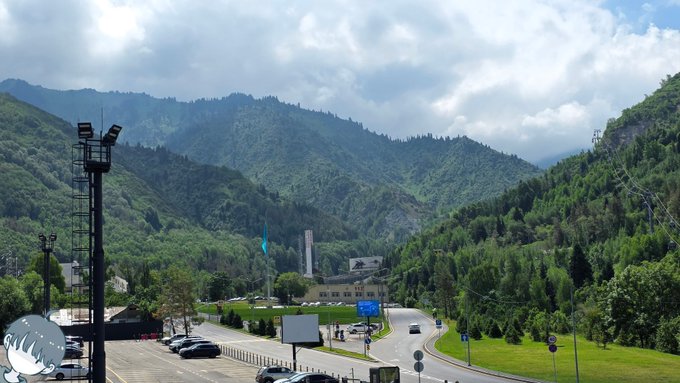 A scenic mountain landscape with lush green forested hills, a winding road, and a parking area in the foreground under a partly cloudy sky.