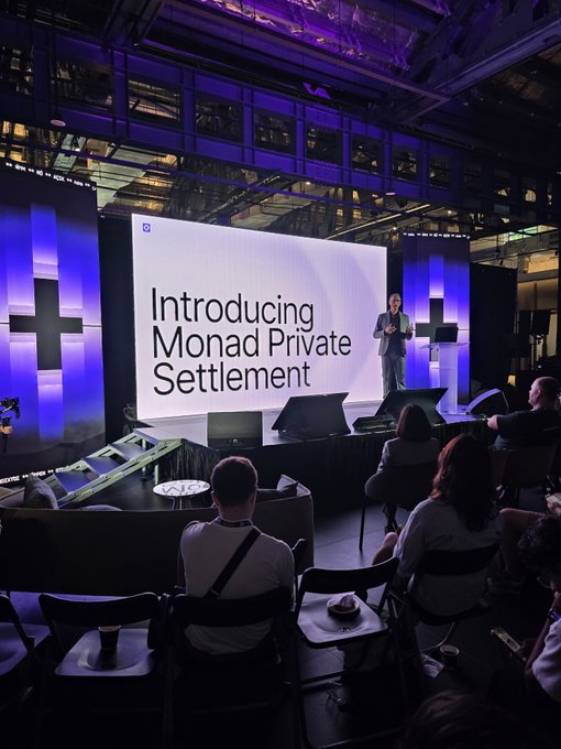 A presentation stage with a large screen displaying 'Introducing Monad Private Settlement,' where a speaker stands addressing an audience seated in chairs in a modern venue with blue lighting.