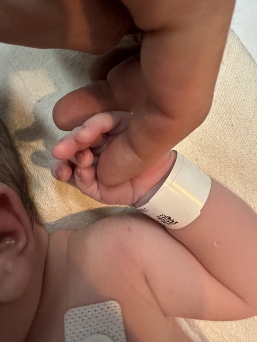 An adult's hand gently holds the tiny fingers of a newborn baby lying on a beige surface, with a hospital identification band visible on the infant's wrist.