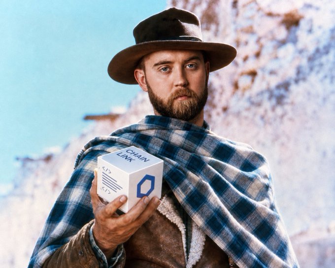 A bearded man in a brown hat and blue plaid poncho holds a white box labeled "CHAIN LINK" with code symbols and a hexagonal logo against a snowy mountain backdrop.