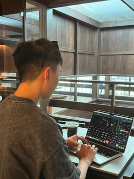 A man with sunglasses perched on his head types on a laptop displaying charts and data, viewed from behind in an indoor setting near a staircase and wooden-paneled walls. The image visually represents the post's scenario of a programmer monitoring an AI trading experiment on Polymarket, capturing the focused, behind-the-scenes moment of letting Claude autonomously execute financial trades.