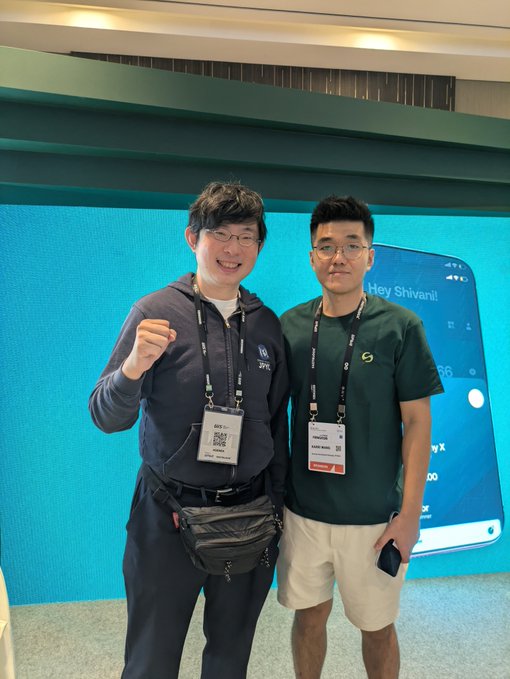Two smiling men wearing lanyards pose for a photo at what appears to be a conference, standing in front of a digital screen displaying 'Hey Shivari!'.
