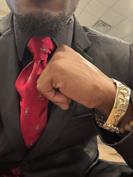 A man in a dark suit and red patterned tie adjusts his collar, showcasing a gold bracelet adorned with diamond-encrusted dollar signs on his wrist. No visible text, numbers, or tickers are present in the image.