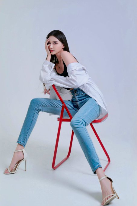 A fashion model with long dark hair poses seated on a red chair against a white backdrop. She wears an oversized white shirt over a black top, blue jeans, and white strappy high heels.