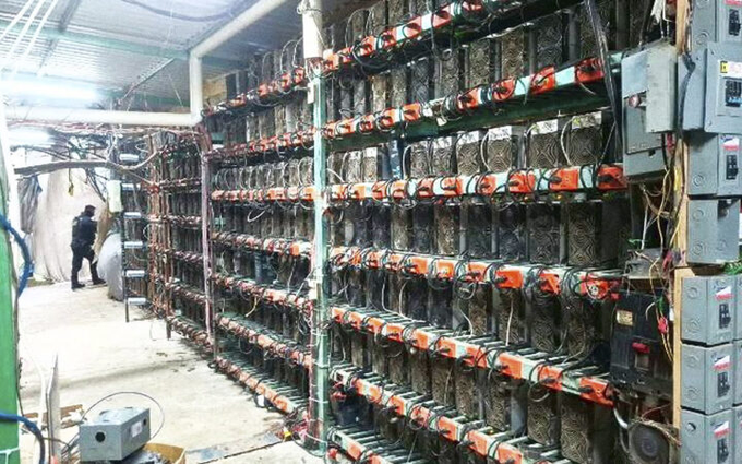 A large warehouse filled with tall racks of cryptocurrency mining hardware, with a person standing in the background among the extensive wiring.