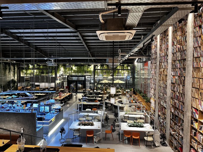 Interior view of a modern, industrial-style office or library space featuring exposed ductwork on the ceiling, desks arranged in rows, and a tall wall of bookshelves on the right.
