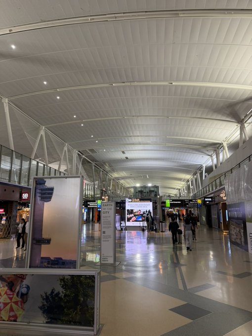 An interior view of an airport terminal or large concourse with high arched ceilings, patterned tile floors, digital advertising displays, and travelers walking in the distance.