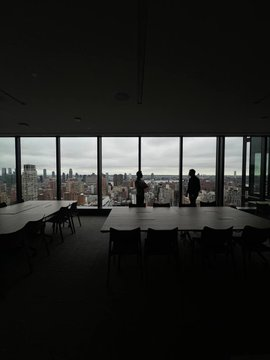 Silhouetted figures overlook a city skyline through floor-to-ceiling windows in a dark conference room with long tables and chairs.