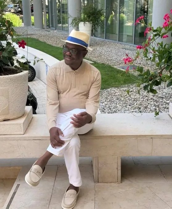 A man in a beige polo shirt, white pants, beige loafers, straw fedora with blue band, and dark sunglasses sits casually on a stone bench outdoors, surrounded by planters with pink bougainvillea flowers and a landscaped courtyard.