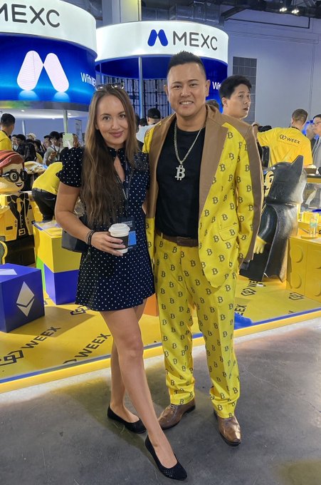A man wearing a yellow patterned suit and a Bitcoin necklace poses with a woman in a navy polka dot dress at a MEXC event, with MEXC and WOO WEEK branding visible in the background.