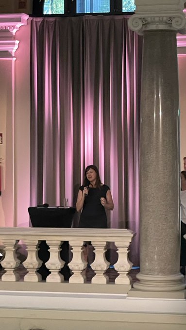 A woman in a black dress speaking into a microphone while standing behind an ornate white marble balustrade, with pink-lit curtains and a large column visible in the elegant venue.