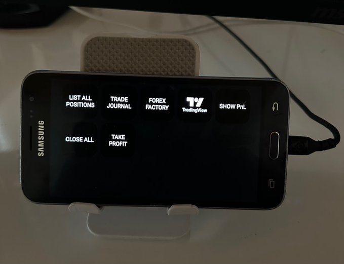 A Samsung smartphone is propped up on a stand, displaying a custom trade terminal interface with buttons for "LIST ALL POSITIONS," "TRADE JOURNAL," "FOREX FACTORY," "TradingView," "SHOW PnL," "CLOSE ALL," and "TAKE PROFIT," illustrating how an old phone was repurposed into a free forex trading dashboard.