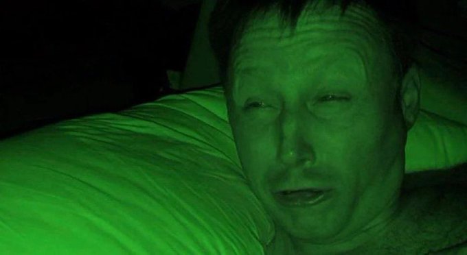 A close-up, green-tinted night-vision style photo of a man lying in bed, appearing sweaty and distressed.