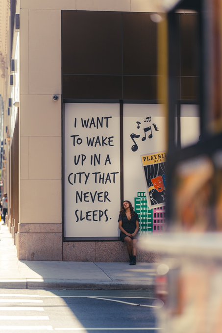 A woman poses in front of a large mural reading 'I WANT TO WAKE UP IN A CITY THAT NEVER SLEEPS,' accompanied by illustrations of musical notes, a Playbill, and city buildings.