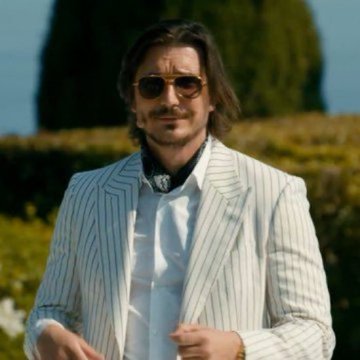 A man with shoulder-length hair, a mustache, and sunglasses wearing a striped white suit jacket and bow tie, standing outdoors near green hedges.