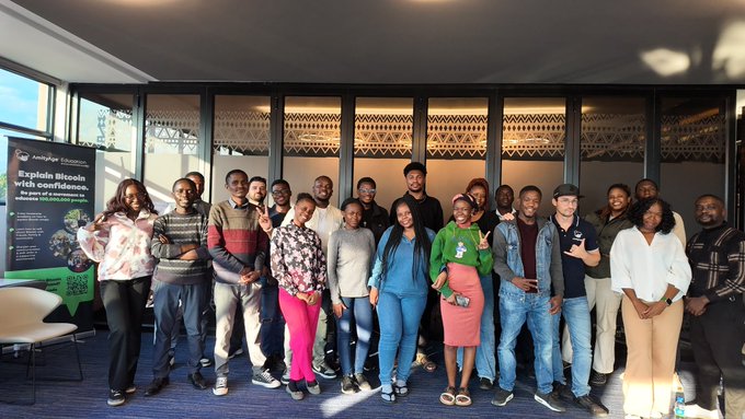 A group of approximately 20 people stands smiling for a photo at what appears to be an 'Antelope Education' event, with a banner promoting 'Explain Bitcoin with confidence.'