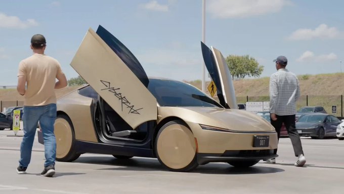 A gold/champagne colored Tesla Cybercab robotaxi with scissor doors open, featuring graffiti reading 'SHARP' on the side and a license plate marked '007', is observed by two people in a parking lot.