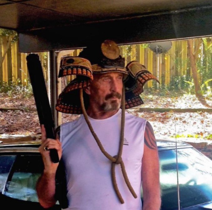 A man wearing a samurai helmet with red cord, a white sleeveless shirt, and holding a shotgun while standing under a carport.