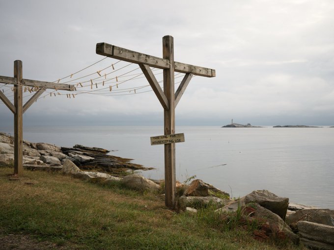 A coastal scene features wooden drying racks with clothespins in the foreground, a rocky shoreline, and a small island with a lighthouse in the distance under an overcast sky.