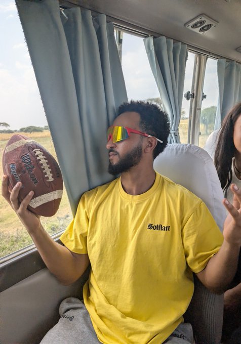 A man wearing a yellow "Solflare" t-shirt and colorful sunglasses holds a Wilson football while sitting on a bus.