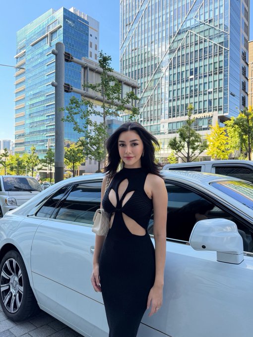 A woman in a black cutout dress poses confidently beside a white luxury sedan, with sleek glass skyscrapers rising in the background. The polished urban setting complements the caption's contrast between high-end city life and the humorous nod to trading losses.