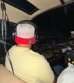 A person wearing a red and white cap and tan jacket sits inside a small aircraft, looking out at a city landscape below.