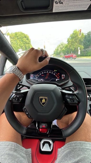 A first-person view from inside a Lamborghini showing a hand on the steering wheel, a luxury watch and ring, with the dashboard visible.