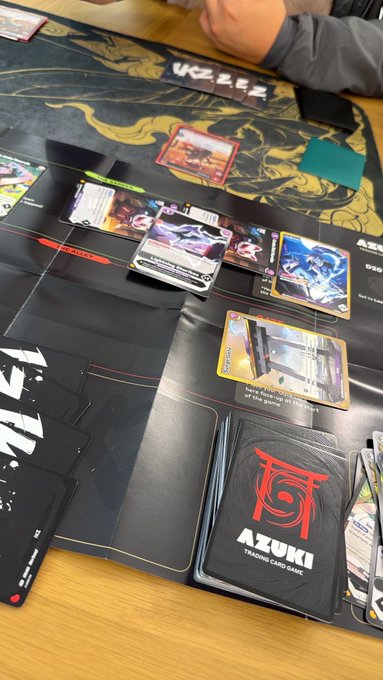 A photo of a Pokémon trading card game in progress on an Azuki playmat, with cards including Cinderace, Greninja, and Iron Hands laid out alongside a deck of cards.