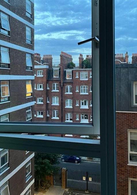A view through a window shows urban brick apartment buildings with chimneys and a parked car below, symbolizing the pursuit of financial success as suggested by "get money (GM)"—representing the hustle of city life and the goal of achieving financial stability.