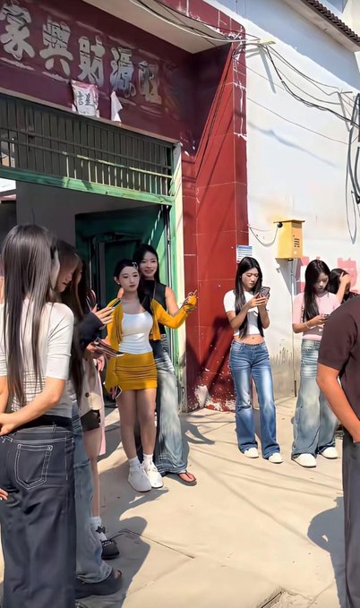 A group of young women, including one in a yellow two-piece outfit holding up a peace sign and others filming on their phones, gather outside a small storefront with Chinese signage on its red facade, illustrating the growing presence of content creators bringing "ethnic flair" to rural village life.