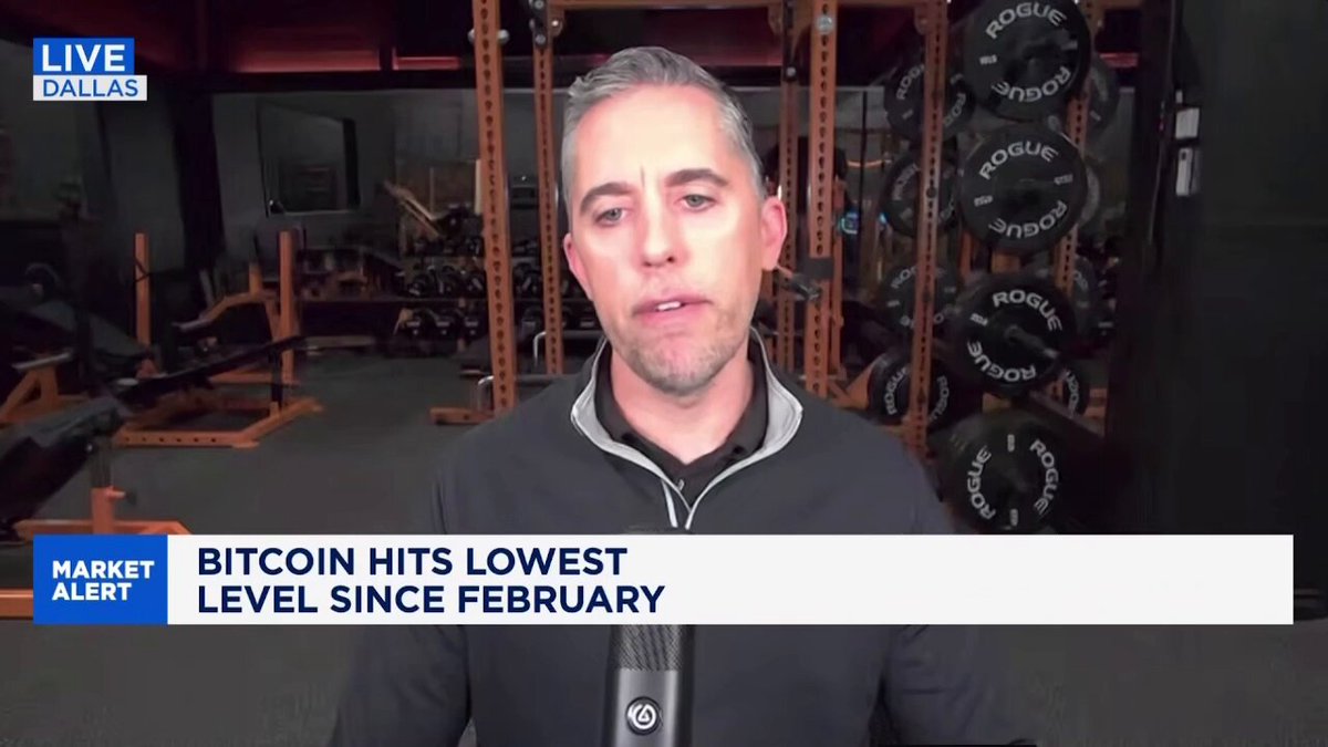 The image features a man wearing a black shirt, standing inside of a gym or fitness center surrounded by exercise equipment like dumbbells and a bench. The man appears to be at work out or speaking about his experiences with fitness and health. A TV screen can be seen in front of him, potentially displaying the news related to cryptocurrency trading and Bitcoin hits. The image seems to focus on the importance of investing in Bitcoin during specific periods due to its current popularity.
