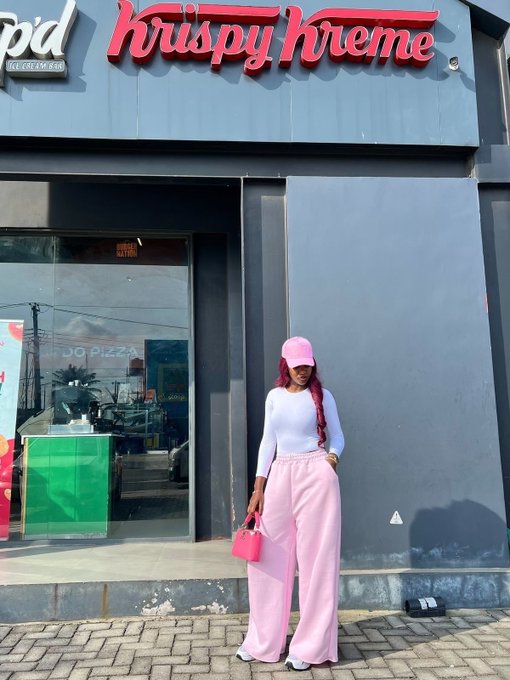 A woman with pink hair wearing a pink cap, white top, and pink pants stands outside a Krispy Kreme shop holding a small pink handbag.