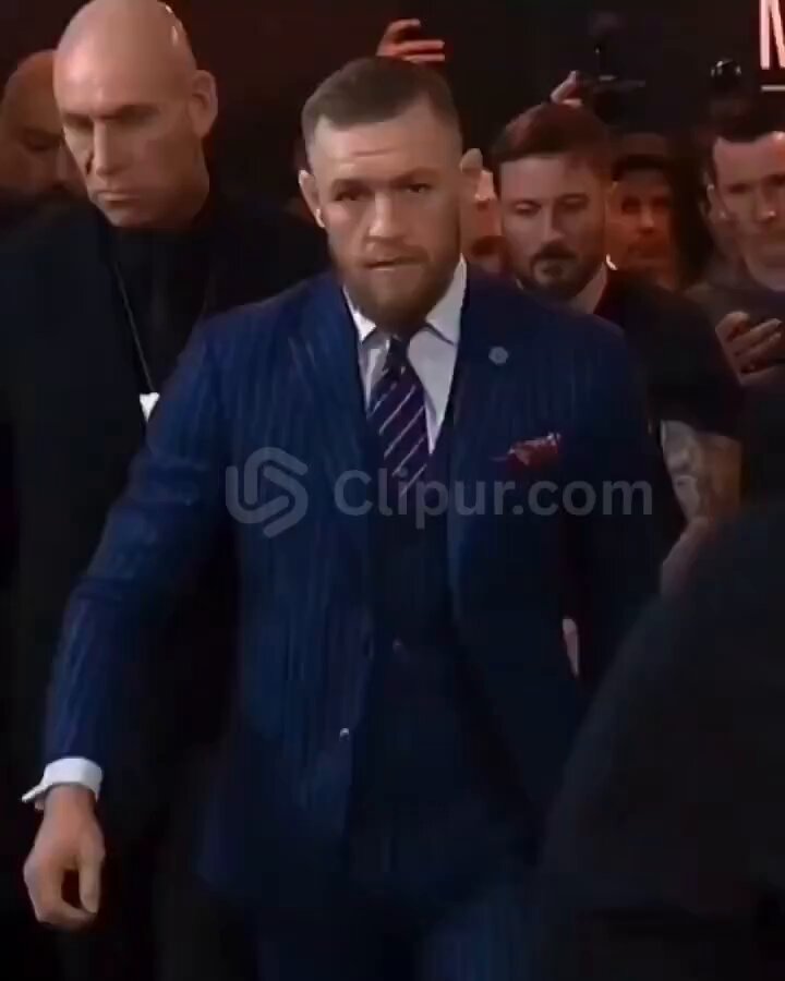 The image features a man in a suit walking down a street, surrounded by other people who seem interested or engaged with him. The man is wearing a striped blue suit and tie, standing out from the crowd. There are numerous people in close proximity to each other, forming a group that appears quite crowded. The overall atmosphere of this scene seems to be quite public and social, likely related to a business event, performance, or special occasion where the man in the suit is an influencer with a large following.