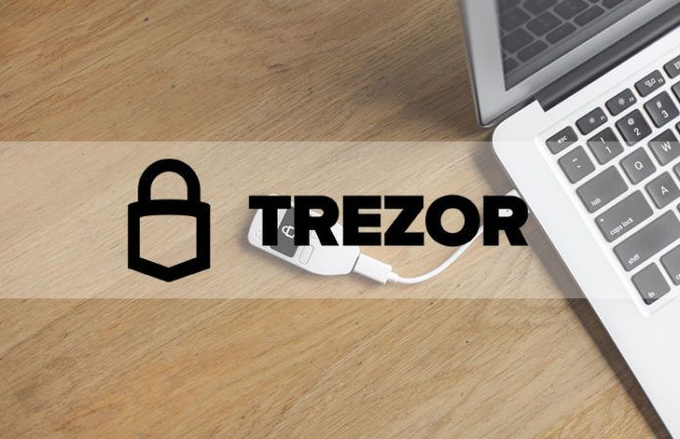 A promotional image showing a Trezor hardware wallet connected via USB to a laptop on a wooden desk, with the Trezor lock logo and brand name displayed.
